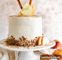 layer apple spice cake with cream cheese frosting garnished with chopped pecans, cinnamon sticks and apple slices on a white cake plate