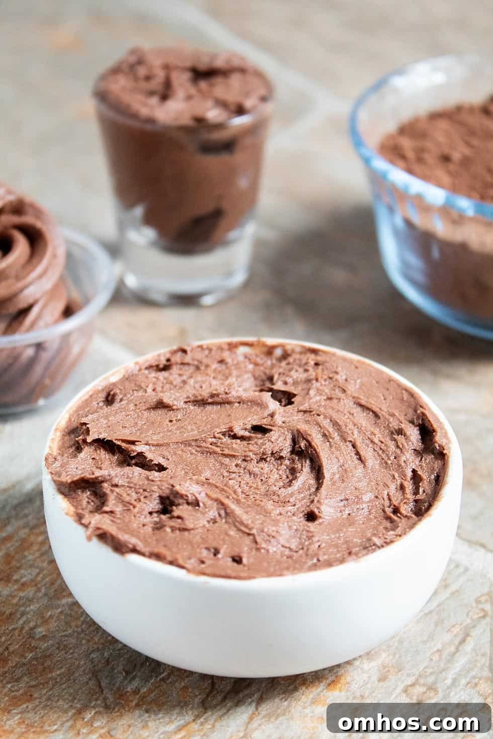 side view of white bowl containing keto chocolate frosting in front of other bowls and cocoa powder