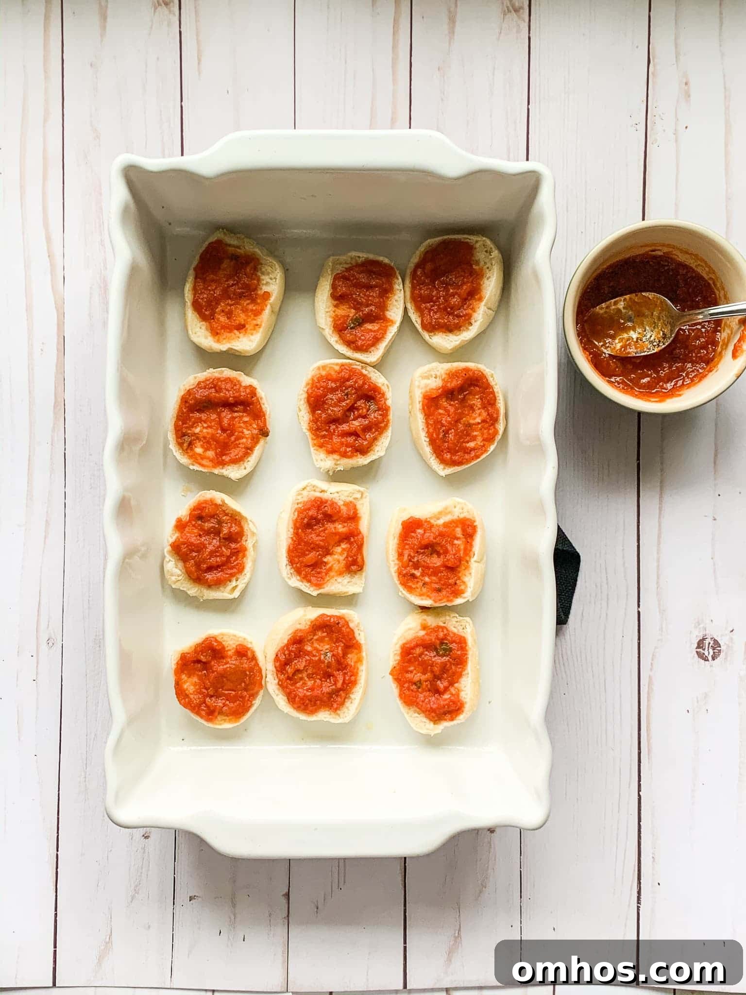 spreading marinara sauce on mini slider buns inside a baking dish.
