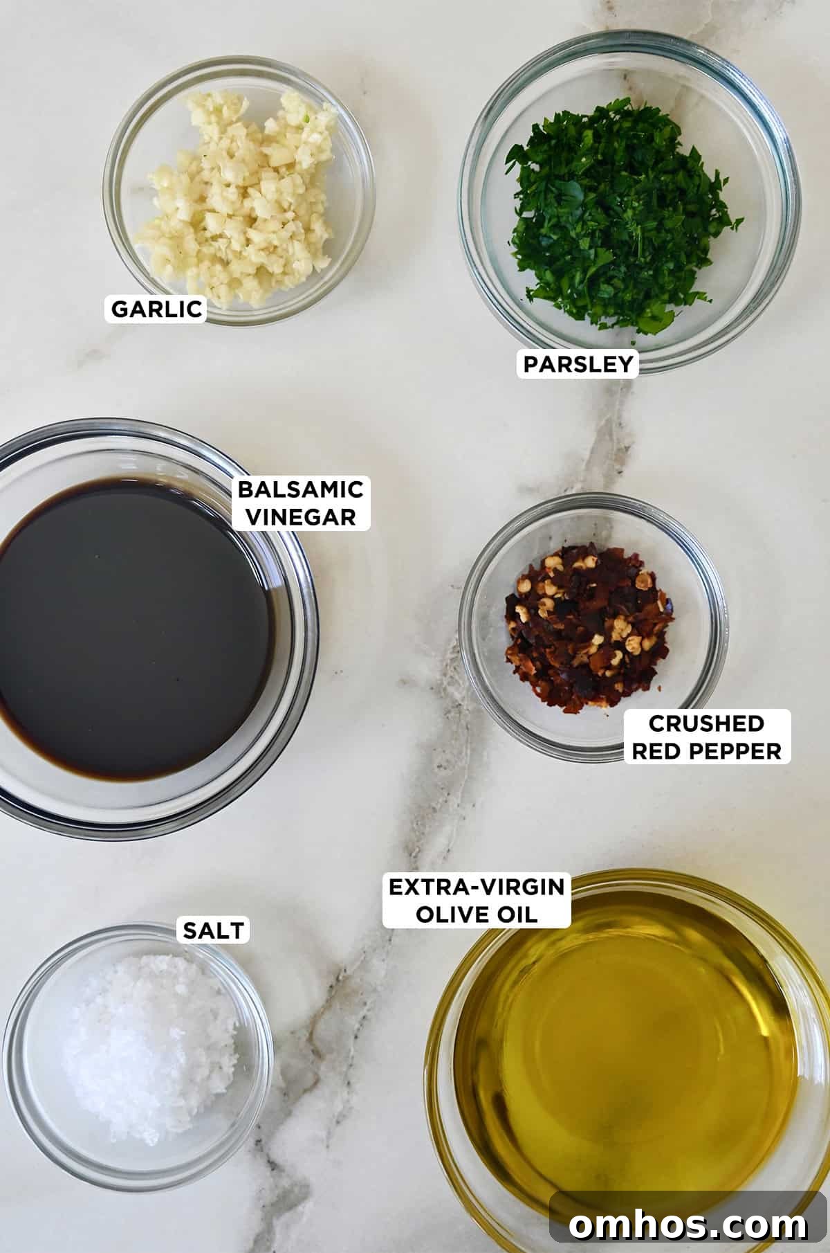 Various sizes of glass bowls containing bread dipping oil ingredients, including minced garlic, parsley, crushed red pepper flakes, extra-virgin olive oil, salt and balsamic vinegar.