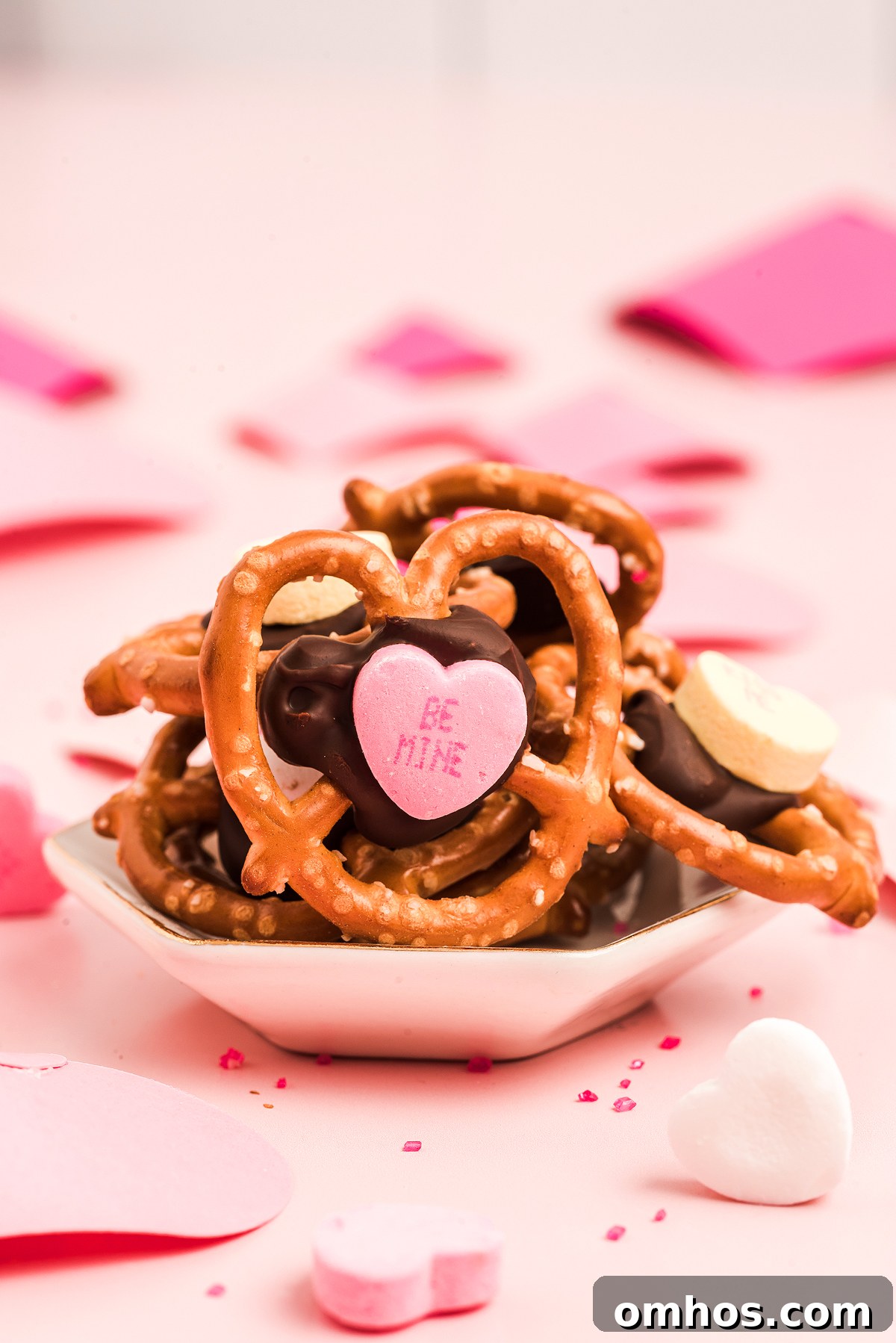 Valentine Pretzel Snack Bites Recipe 11 Heart-shaped Valentine pretzel snack bites with chocolate and pink candy hearts on a small dish, surrounded by festive heart decorations—an adorable Valentine’s Day treat.