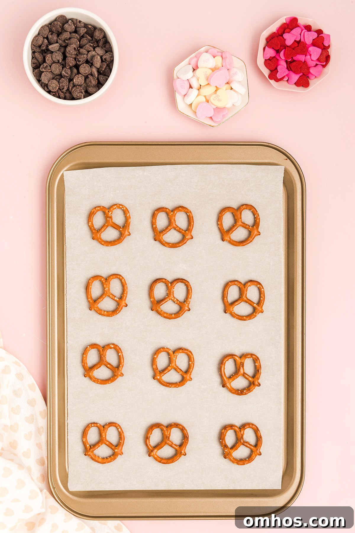 Valentine Pretzel Snack Bites Recipe 13 A baking sheet with Valentine Pretzel Snack Bites, surrounded by bowls of chocolate chips and heart-shaped candies on a pink background.