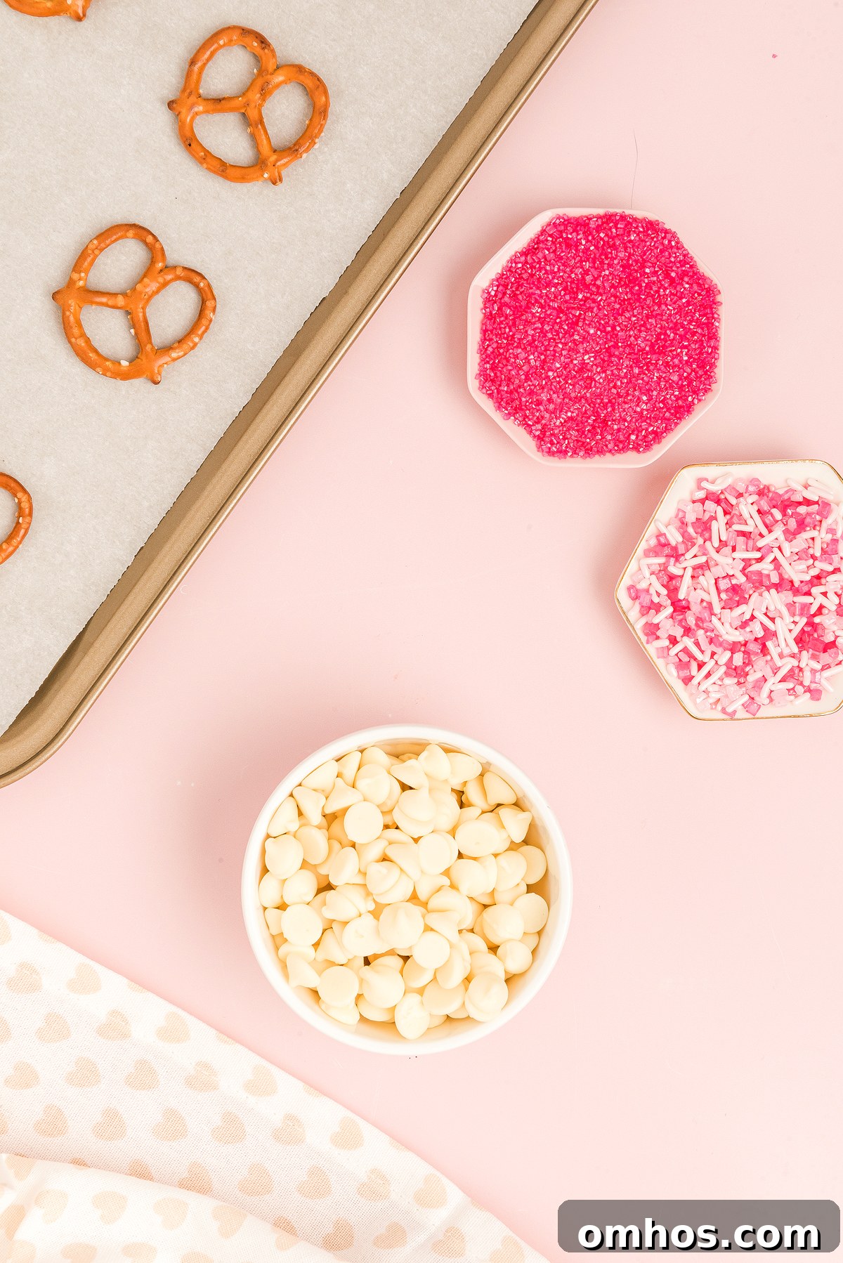 Valentine Pretzel Snack Bites Recipe 14 Baking tray with Valentine Pretzel Snack Bites, a bowl of white chocolate chips, and dishes of pink sprinkles and shredded coconut—perfect for a festive Valentine’s Day treat.
