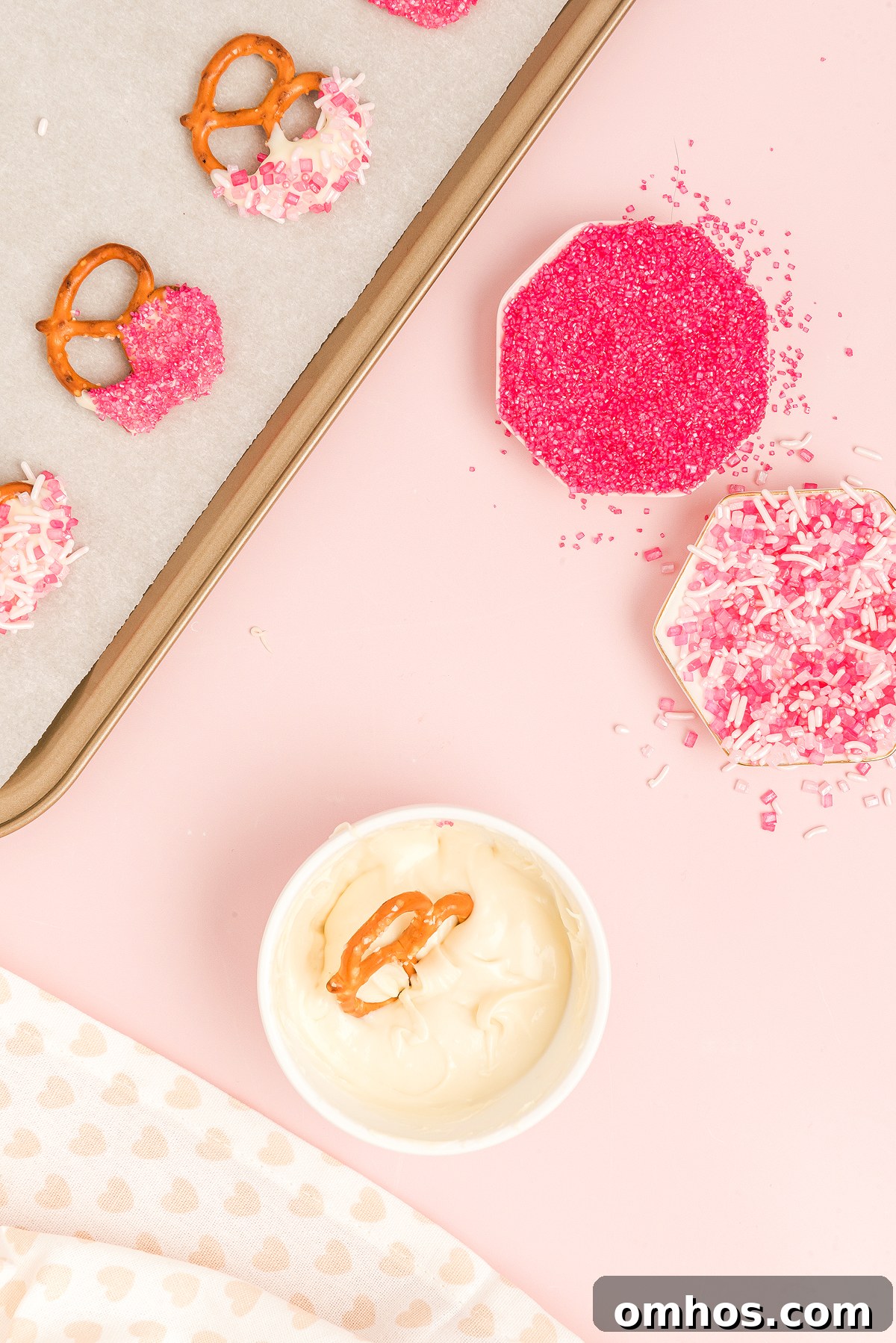 Valentine Pretzel Snack Bites Recipe 15 Pretzels with pink sprinkles and white dip on a pink surface make delightful Valentine Pretzel Snack Bites, presented with a baking tray and small dishes.