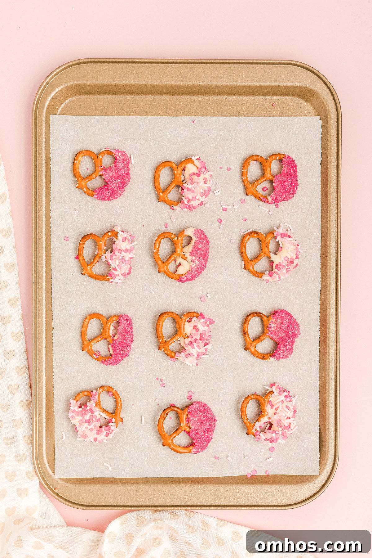 Valentine Pretzel Snack Bites Recipe 16 Baking sheet with Valentine pretzels dipped in white chocolate and pink sprinkles on parchment paper, set against a pink background—a sweet Valentine’s Day treat.