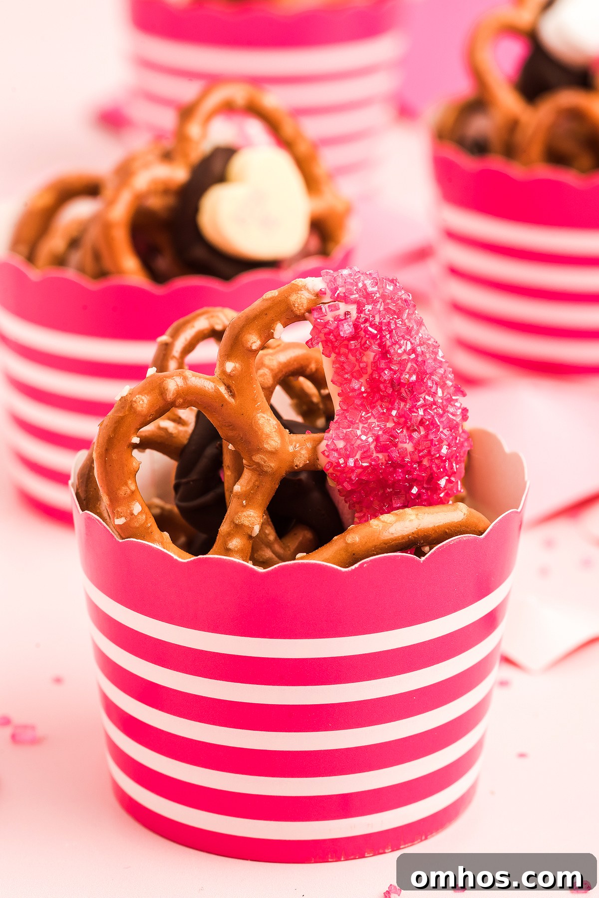 Valentine Pretzel Snack Bites Recipe 6 Close-up of Valentine pretzels and chocolates in a pink-striped cup, topped with a pink sugar-coated treat—perfect as a Valentine’s Day treat.