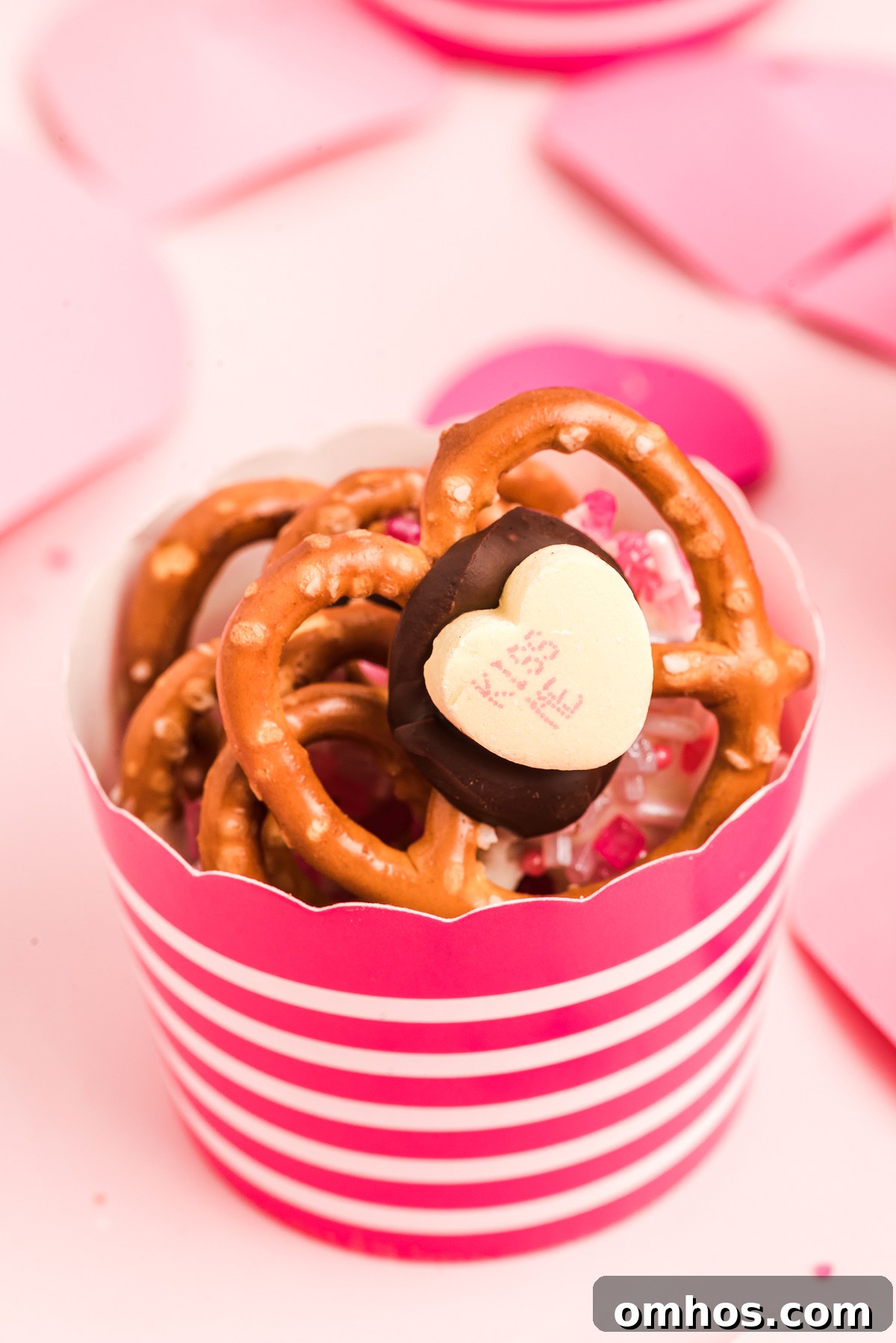 Valentine Pretzel Snack Bites Recipe 7 A cup with striped pink paper holds Valentine Pretzel Snack Bites—pretzels topped with chocolate and a heart-shaped candy, perfect as a Valentine’s Day treat.