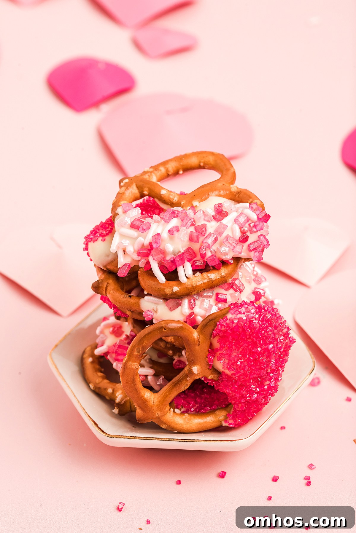Valentine Pretzel Snack Bites Recipe 8 Stack of Valentine pretzels with white chocolate and pink sprinkles on a small plate, with pink paper hearts in the background—a perfect Valentine’s Day treat.