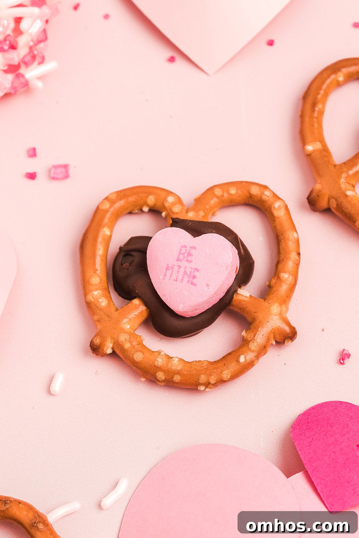 Valentine Pretzel Snack Bites Recipe 10 A Valentine Pretzel Snack Bite topped with chocolate and a pink candy heart that says "BE MINE" sits on a pink background—a perfect Valentine’s Day treat.