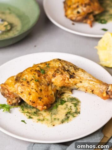 A serving of golden brown roasted chicken leg quarters with an herb sauce on a gray table with more chicken in the background.