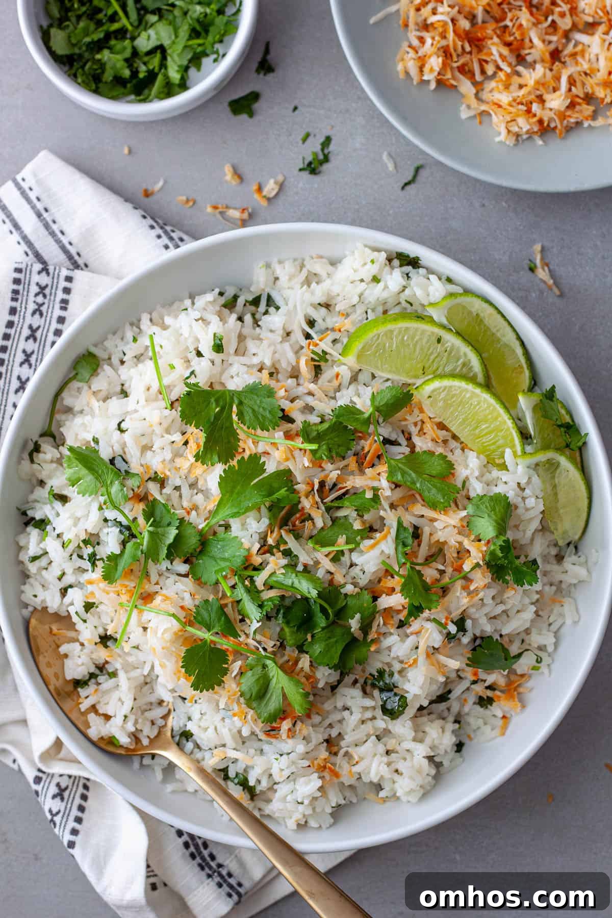 Fragrant Coconut Cilantro Rice Recipe 9 A white serving bowl with coconut cilantro rice topped with toasted shredded coconut and cilantro leaves with lime wedges to the side.