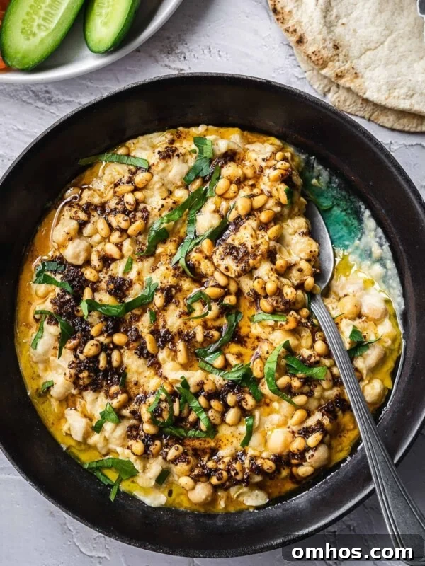 Bowl of warm chickpeas and tahini, garnished with parsley and pine nuts served with sliced cucumbers and pita.