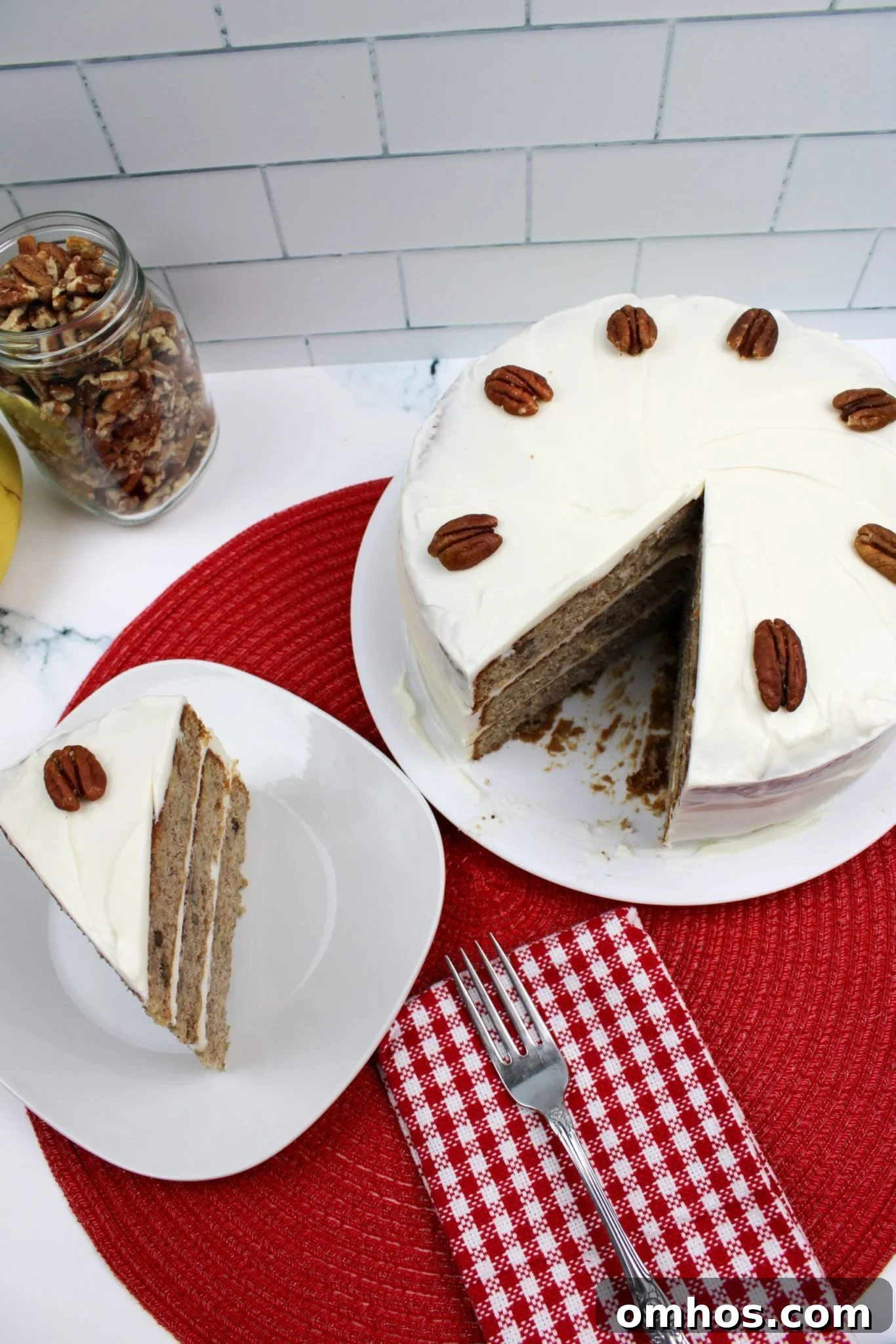 Old-Fashioned Hummingbird Cake Recipe 2 A sliced hummingbird cake with cream cheese frosting and whole pecans on top, displayed on a white plate with a red woven placemat.