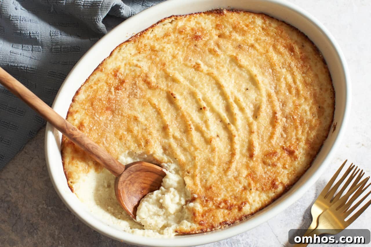 An oval gratin dish of goat cheese mashed potatoes with a wooden spoon in it. A blue towel is in the upper left corner, two gold forks are on the bottom right.