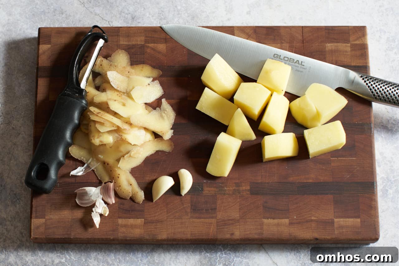 Cubed potatoes, garlic cloves, potato peels, a vegetable peeler, and a chef's knife on a cutting board.