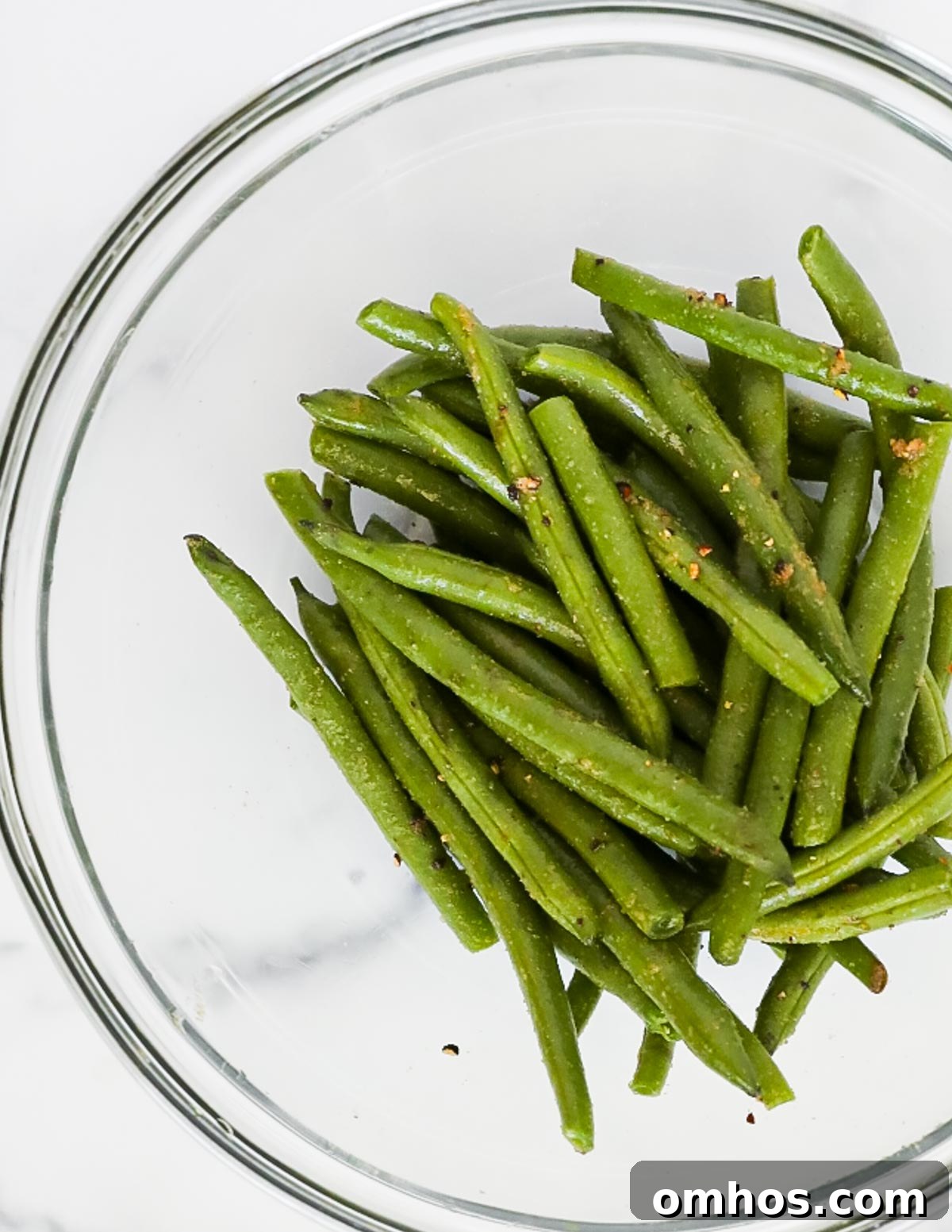 A clear glass bowl filled with fresh green beans covered in olive oil and seasonings.
