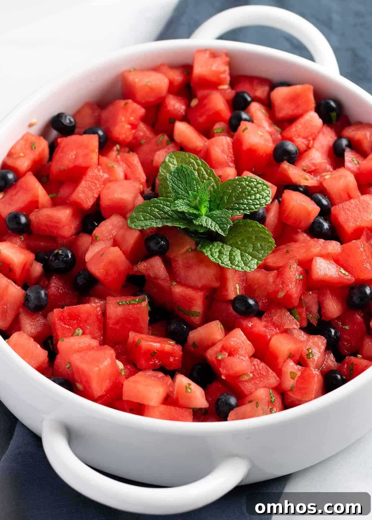 A white serving platter on a blue cloth napkin, filled with fresh watermelon, blueberries and a spring of mint on top. 