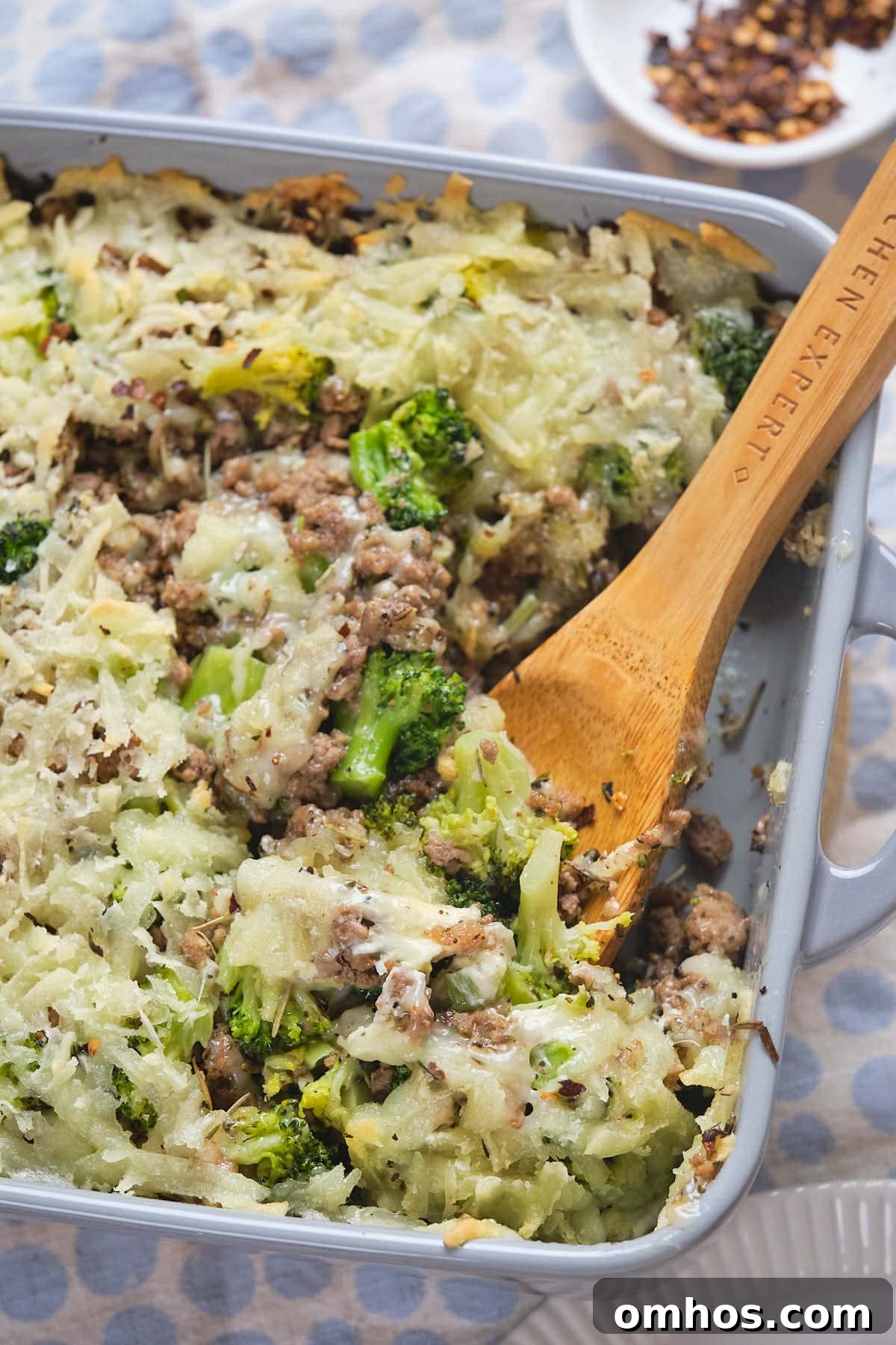 A casserole dish with a wooden spoon, featuring a mixture of cooked ground meat, broccoli, and melted cheese.