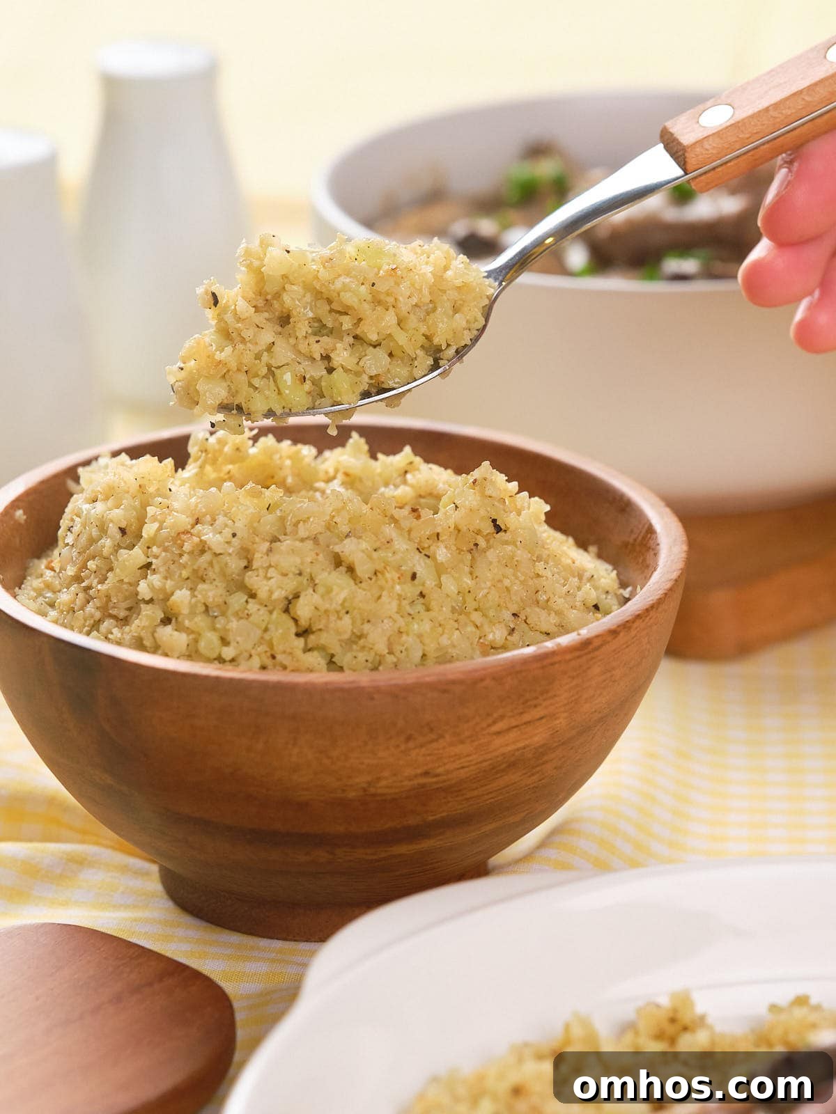 A hand holds a spoonful of cooked cauliflower rice above a wooden bowl filled with more cauliflower rice.