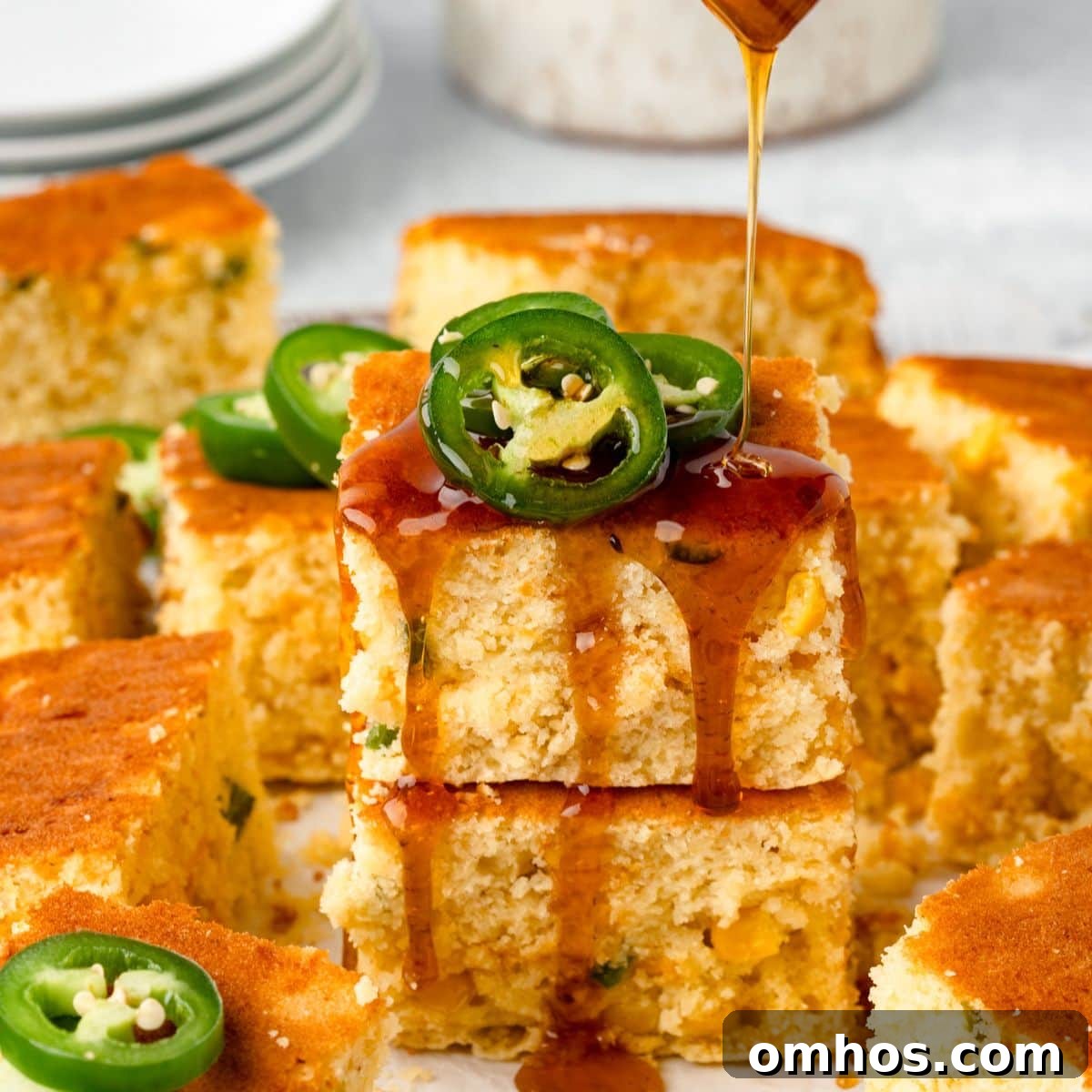 Skillet Jalapeño Cornbread Recipe 3 A stack of cornbread squares topped with sliced jalapeños, drizzled with syrup. The golden-brown cornbread is moist with visible corn kernels. More cornbread pieces and a stack of plates are in the background.