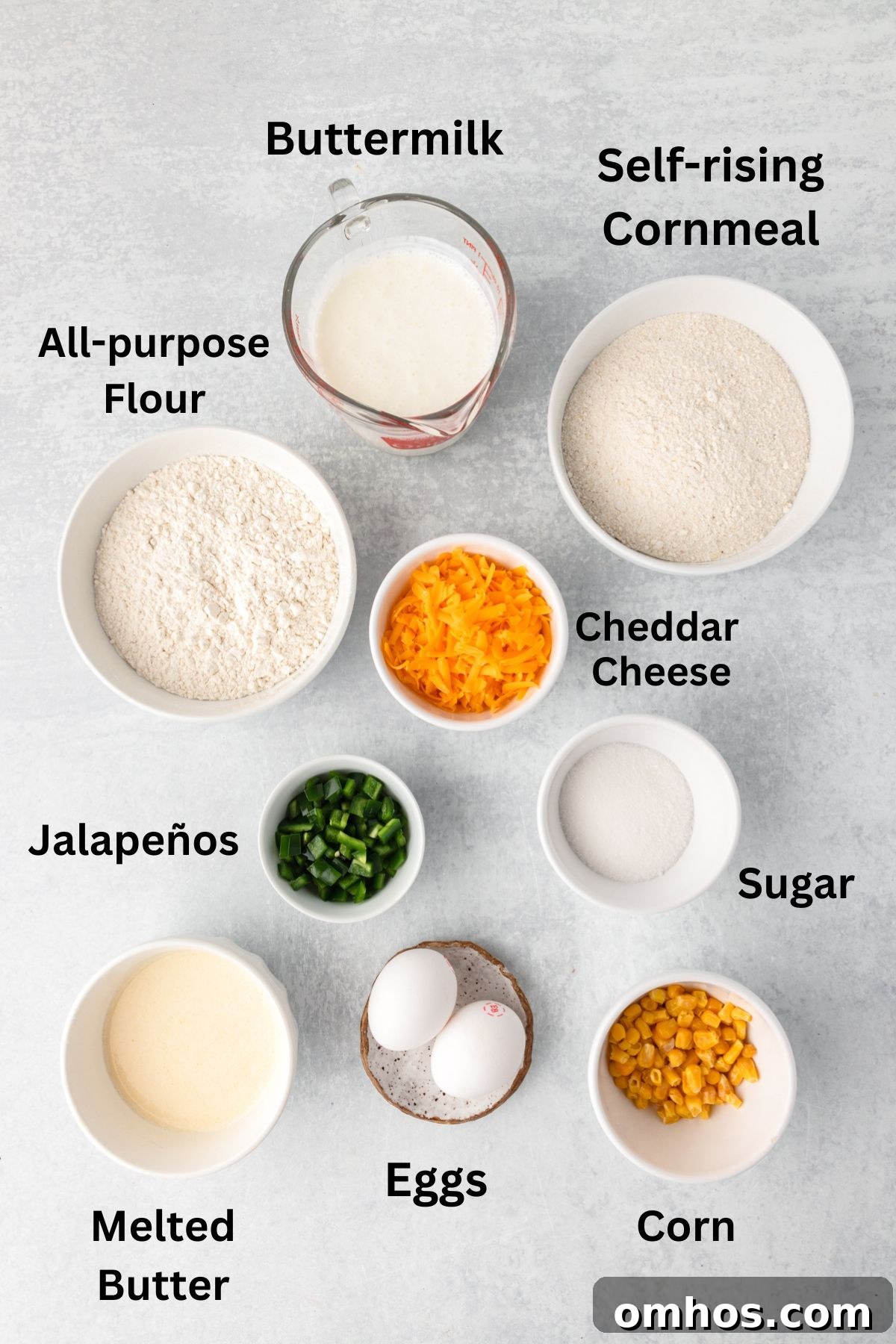 Skillet Jalapeño Cornbread Recipe 4 A top view of cornbread ingredients on a gray surface, including labeled bowls of buttermilk, self-rising cornmeal, all-purpose flour, cheddar cheese, jalapeños, sugar, corn, melted butter, and two eggs.