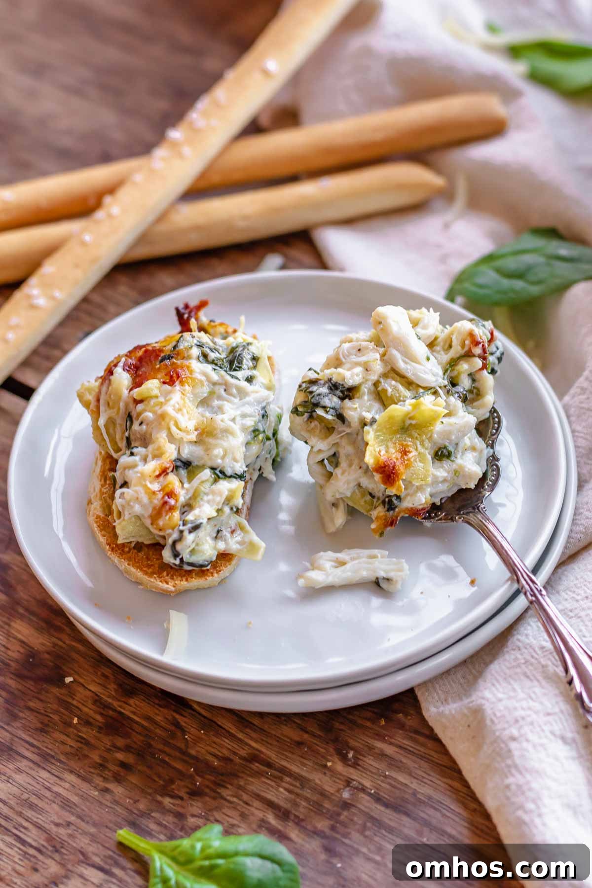 A spoonful of spinach artichoke crab dip on a baguette and some on a spoon resting on a plate.