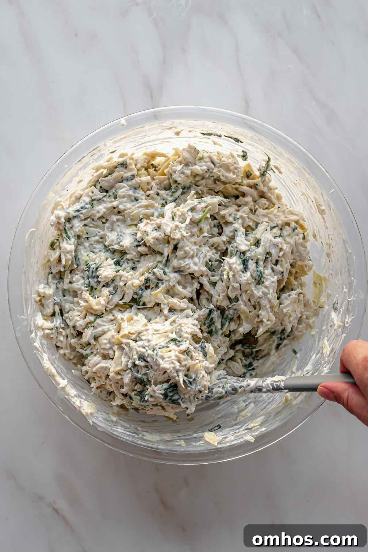 A rubber spatula folds crab meat into spinach artichoke dip.