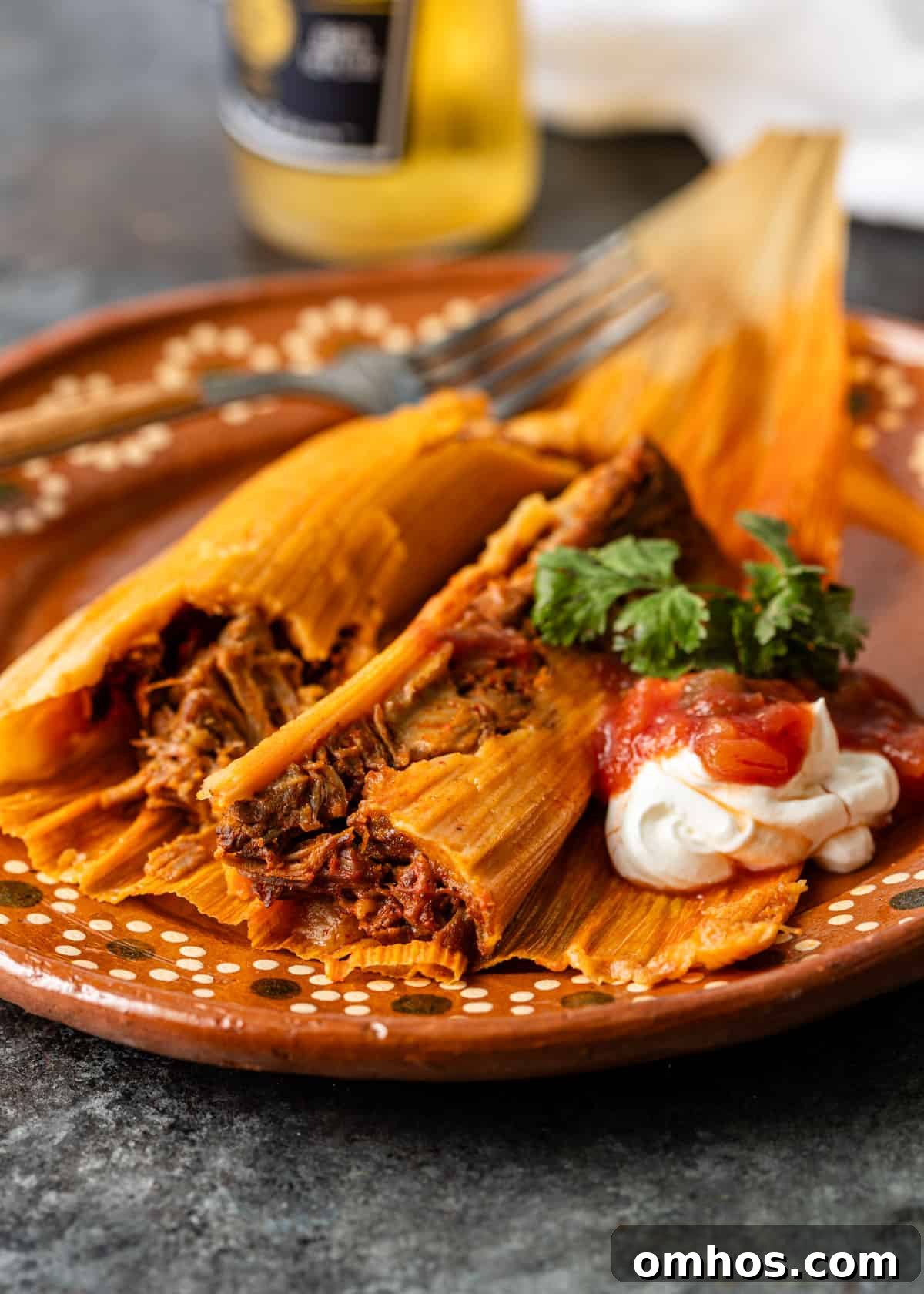 Authentic Mexican Red Pork Tamales Recipe 19 closeup side view: pork tamales on an orange ceramic plate with sour cream, salsa, and fresh herbs on top
